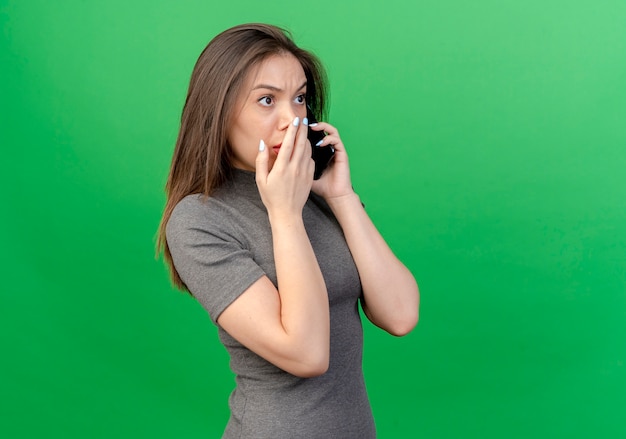 Foto gratuita mujer bonita joven sorprendida de pie en la vista de perfil mirando directamente poniendo la mano en la boca aislada sobre fondo verde con espacio de copia