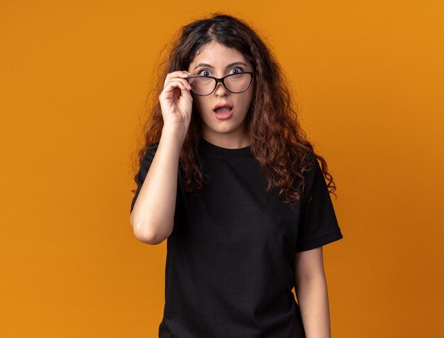 Mujer bonita joven sorprendida vistiendo y agarrando gafas mirando al frente aislado en la pared naranja con espacio de copia