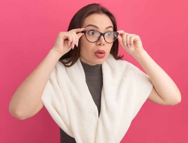 Mujer bonita joven sorprendida vistiendo y agarrando gafas mirando al lado aislado en la pared rosa