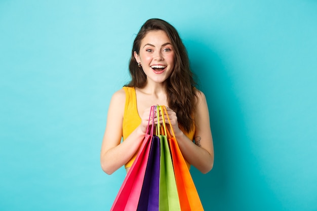 Mujer bonita joven sosteniendo bolsas de la compra, sonriendo emocionado a la cámara, comprando con descuentos, de pie sobre fondo azul.