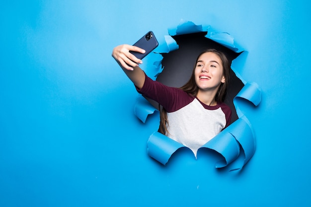 La mujer bonita joven toma el selfie en el teléfono mientras que mira a través del agujero azul en la pared de papel.