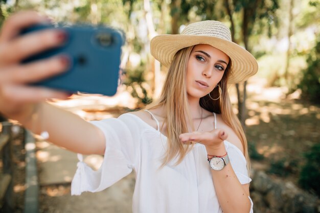 Mujer bonita joven tomando un selfie con un elegante sombrero de verano en un parque.