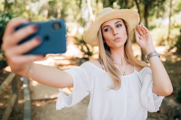 Mujer bonita joven tomando un selfie con un elegante sombrero de verano en un parque.