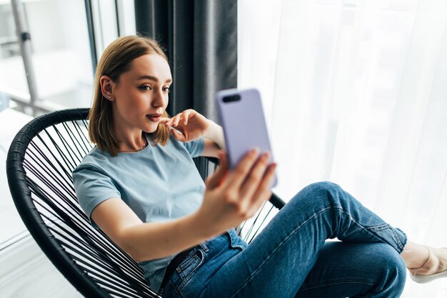 Mujer bonita joven tomando selfie junto a la ventana con persianas en casa