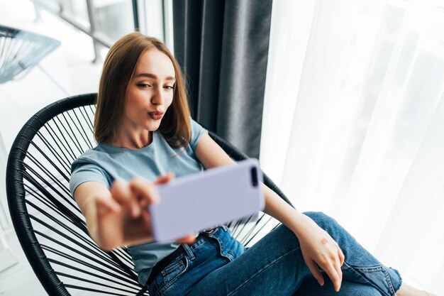 Mujer bonita joven tomando selfie junto a la ventana con persianas en casa