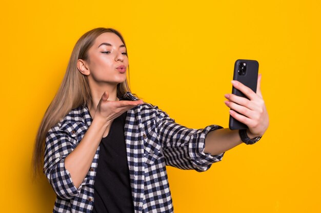 Mujer bonita joven tomar selfie en el teléfono golpe beso aislado en la pared amarilla