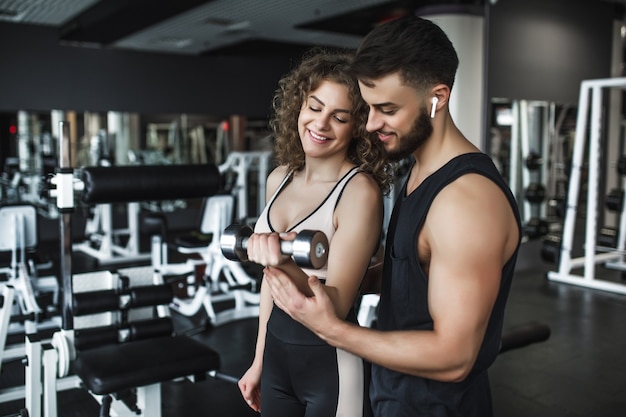 Mujer bonita joven trabajando en el gimnasio, haciendo flexiones de bíceps con la ayuda de su entrenador personal