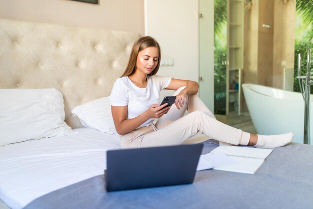 Mujer bonita joven usa el teléfono y la computadora portátil sentada en la cama