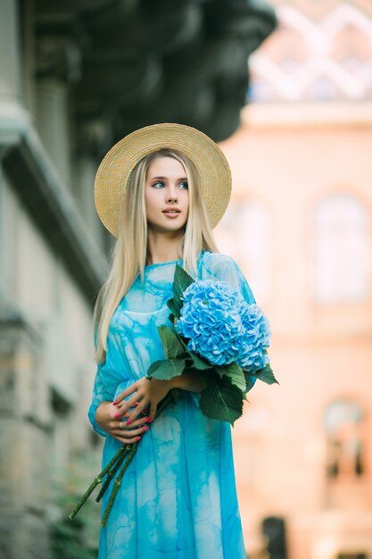 Mujer bonita joven en vestido azul sosteniendo las flores de hydragea en la calle de verano