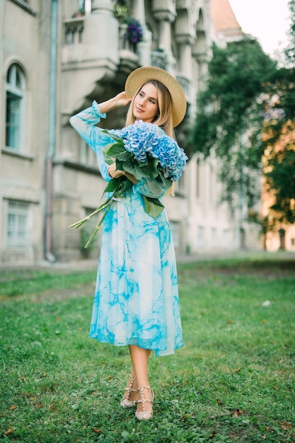 Mujer bonita joven en vestido azul sosteniendo las flores de hydragea en la calle de verano