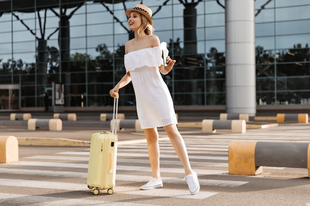 Mujer bonita joven en vestido blanco de gran tamaño mira en la distancia, sostiene la maleta amarilla y el pasaporte