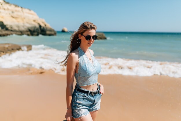 Mujer bonita joven en vestido y sombrero de paja caminando en la playa del océano