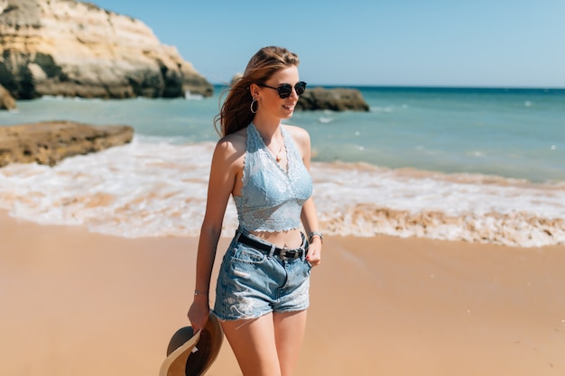 Mujer bonita joven en vestido y sombrero de paja caminando en la playa del océano