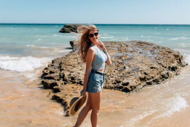 Mujer bonita joven en vestido y sombrero de paja caminando en la playa del océano