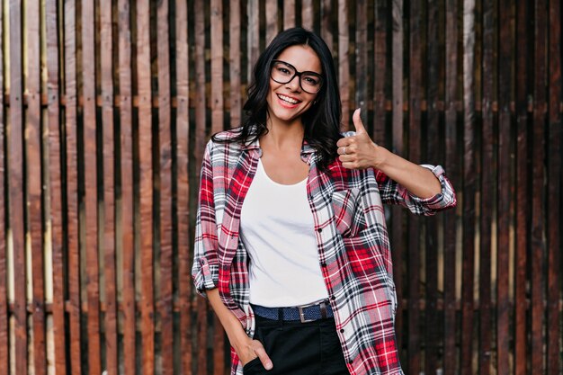 Mujer bonita latina posando con el pulgar hacia arriba en la pared de madera. Foto al aire libre de una chica encantadora relajada lleva gafas y cinturón de cuero.