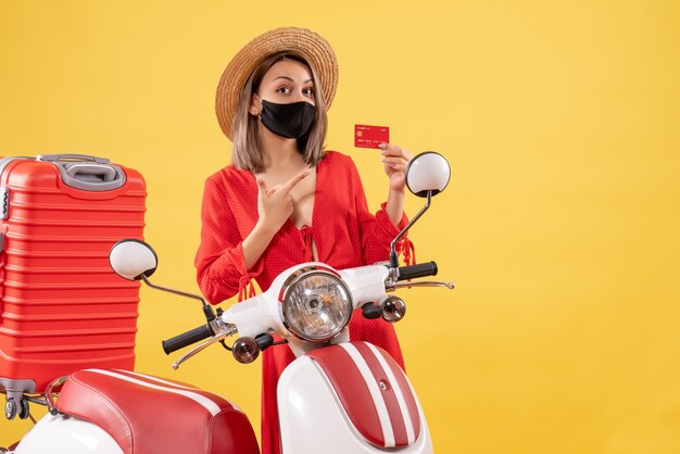 Mujer bonita con máscara negra con tarjeta de crédito de pie cerca de ciclomotor