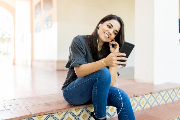 Mujer bonita de mediana edad que escucha la transmisión de radio a través de una aplicación móvil mientras está sentada en la entrada