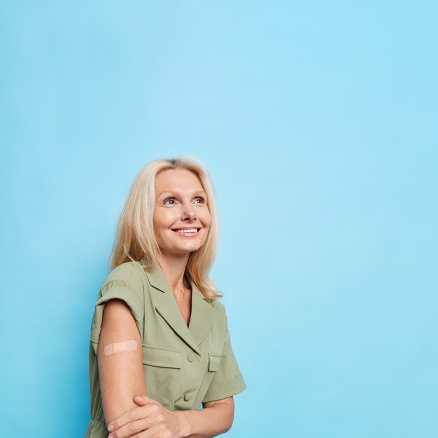 Mujer bonita de mediana edad rubia complacida pensativa usa vendaje adhesivo en el brazo feliz después de hacer la vacunación enfocada sobre poses contra la pared azul