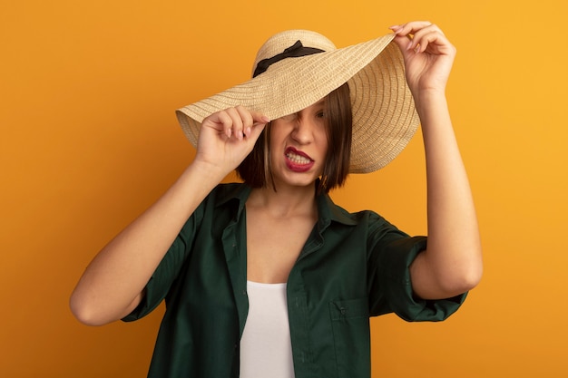 Foto gratuita mujer bonita molesta cubre la cara con sombrero de playa aislado en la pared naranja