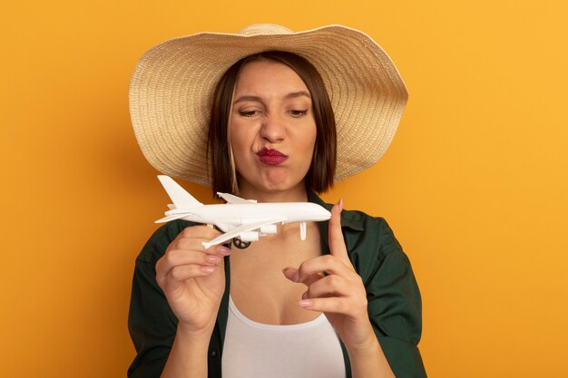 Mujer bonita molesta con sombrero de playa sostiene y mira el modelo de avión aislado en la pared naranja