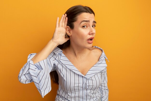 Mujer bonita molesta tiene la mano detrás de la oreja tratando de escuchar aislado en la pared naranja