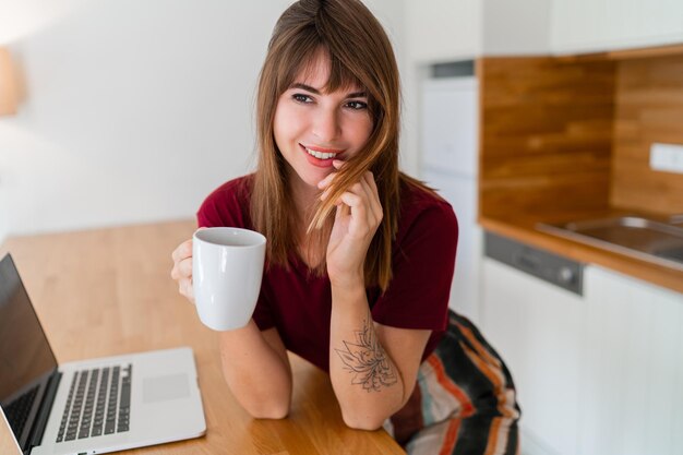 Mujer bonita morena bebiendo café y sentada en la cocina moderna y usando una computadora portátil Buscando trabajo en línea