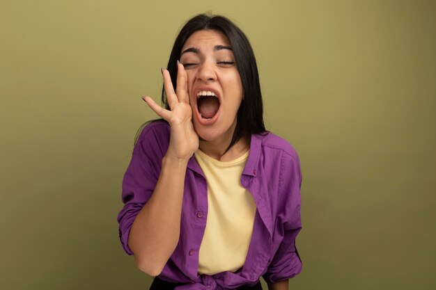 Mujer bonita morena disgustada sostiene la mano cerca de la boca llamando a alguien aislado en la pared verde oliva