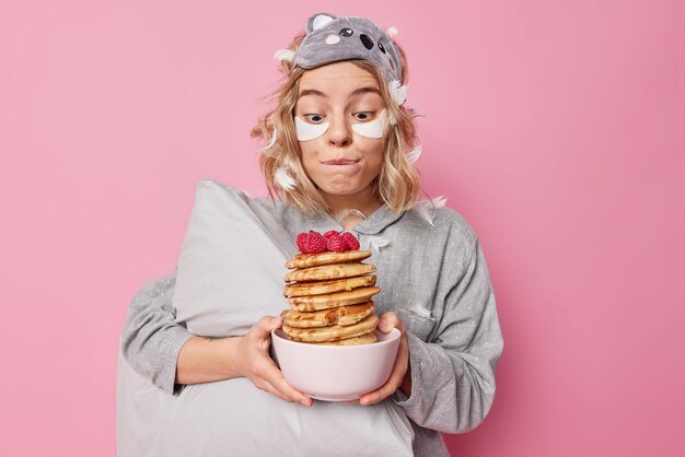 Mujer bonita muerde los labios mira deliciosos panqueques con frambuesa va a comer un sabroso desayuno apetitoso vestido con ropa de dormir sostiene la almohada aplica parches de belleza posa en el interior contra la pared rosa