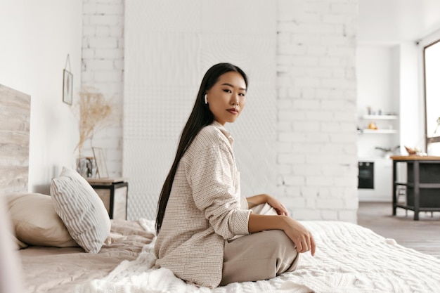 Una mujer bonita con pantalones beige y cárdigan mira a la cámara, medita, escucha música relajante con auriculares inalámbricos y se sienta en una cama blanda