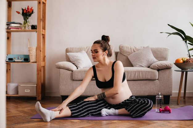 Mujer bonita en pantalones de rayas y top negro estirando y escuchando música en auriculares. Chica embarazada de buen humor haciendo yoga en la alfombra púrpura.