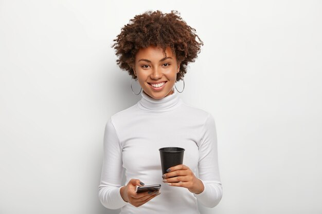 Mujer bonita con peinado afro, tiene un teléfono móvil moderno y café para llevar, pasa tiempo libre charlando en línea