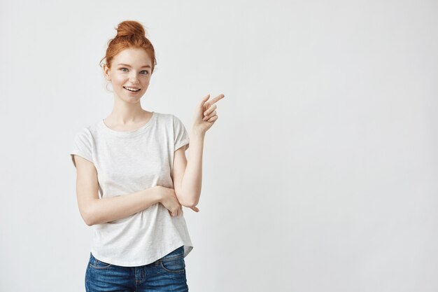 Mujer bonita pelirroja con pecas sonriendo apuntando el dedo a la derecha.