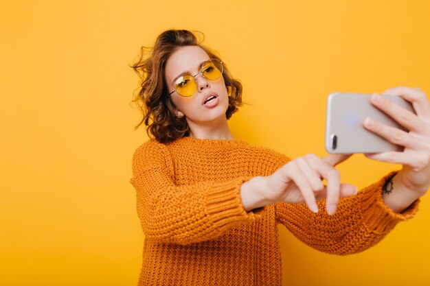 Mujer bonita con pelo corto y rizado sosteniendo smartphone y escribiendo mensaje frente a pared amarilla