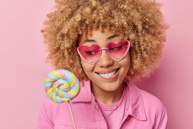 Una mujer bonita con el pelo rizado muerde los labios sostiene una piruleta redonda tiene un diente dulce usa gafas de sol de moda y una chaqueta aislada sobre fondo rosa Modelo femenino positivo mira deliciosos dulces