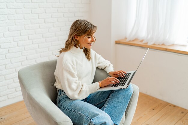 Mujer bonita con pelos ondulados rubios sentado en el sofá en casa trabajando en la computadora portátil