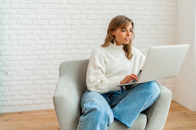 Mujer bonita con pelos ondulados rubios sentado en el sofá en casa trabajando en la computadora portátil