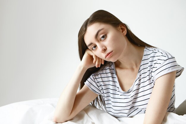 Mujer bonita en pijama de rayas habiendo aburrido expresión infeliz, mirando con la mano debajo de la mejilla mientras está sentado en la cama