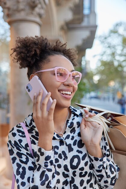 Una mujer bonita positiva con moños de pelo rizado hace una llamada lleva bolsas de compras se ve feliz vestida con ropa de moda pasea al aire libre contenta de comprar ropa nueva en la boutique Consumismo