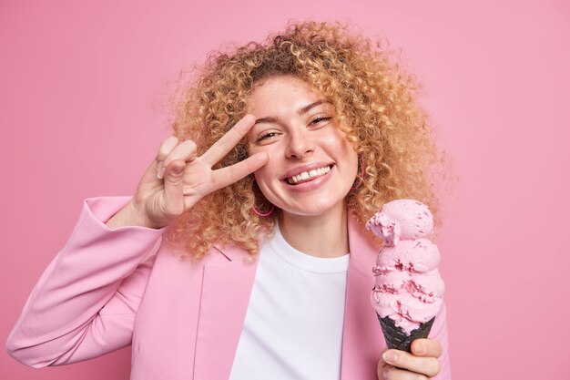 Una mujer bonita positiva con el pelo rizado come un helado enorme en un gofre de cono hace que el gesto de la paz sonríe ampliamente se divierte durante el día de verano vestida con ropa elegante aislada sobre una pared rosa