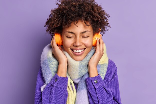 Una mujer bonita y rizada disfruta escuchando música agradable, mantiene los ojos cerrados, usa auriculares estéreo con sonido de buena calidad, usa una elegante chaqueta con bufanda alrededor del cuello.