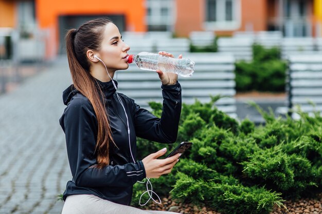 Mujer bonita en ropa deportiva, agua potable en el aire fresco después de correr por la mañana. Concepto saludable.