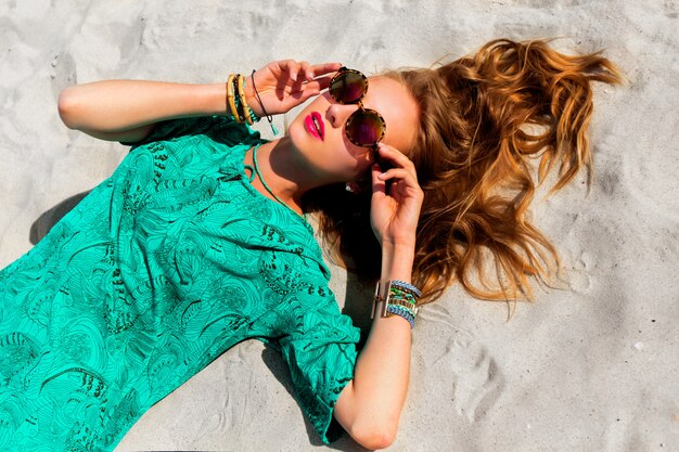 Mujer bonita rubia acostada en la playa tropical soleada, con gafas de sol con estilo fresco, túnica boho de color y accesorios de moda brillantes