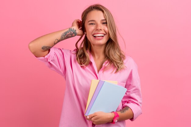 Mujer bonita rubia en camisa rosa sonriendo sosteniendo cuadernos de explotación