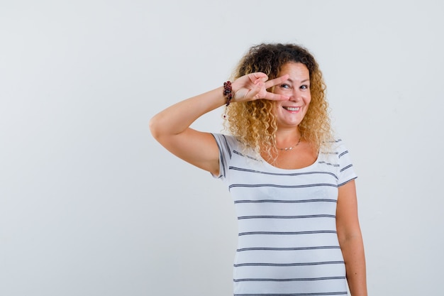 Mujer bonita rubia en camiseta a rayas que muestra el signo V en el ojo y mirando alegre, vista frontal.