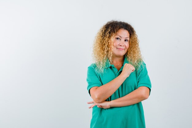 Mujer bonita rubia manteniendo el brazo en el pecho en camiseta de polo verde y mirando alegre. vista frontal.