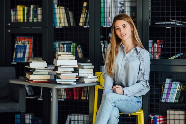 Mujer bonita rubia o modelo sentada en la biblioteca de la universidad con libros sobre la mesa, sosteniendo vasos en las manos