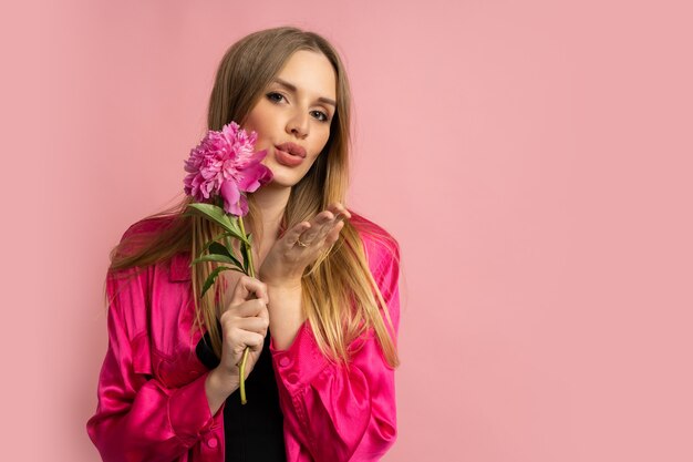 Mujer bonita rubia posando con flor de peonía en elegante traje de verano sobre pared rosa