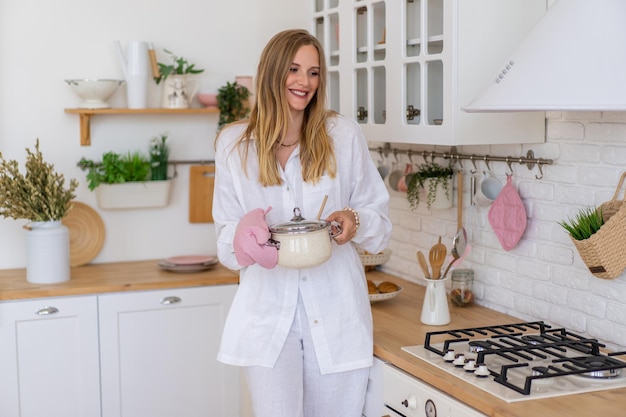 Mujer bonita rubia vestida con traje de lino blanco preparar comida en su cocina, concepto de ama de casa perfecta.