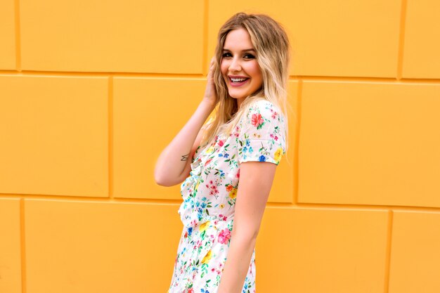 Mujer bonita rubia con vestido floral, posando junto a la pared amarilla