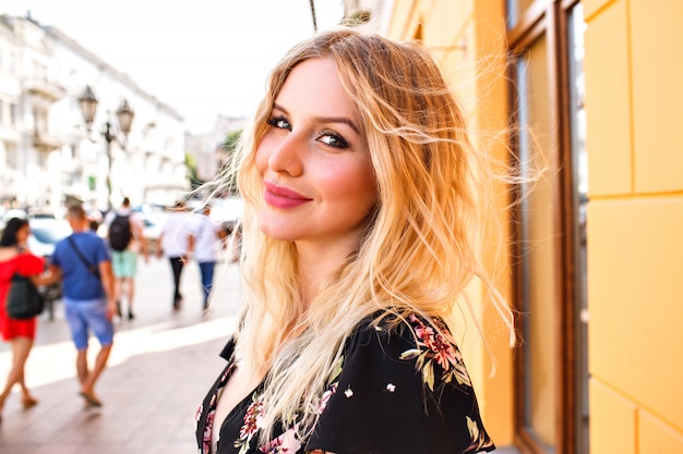 Mujer bonita rubia con vestido floral, posando junto a la pared amarilla